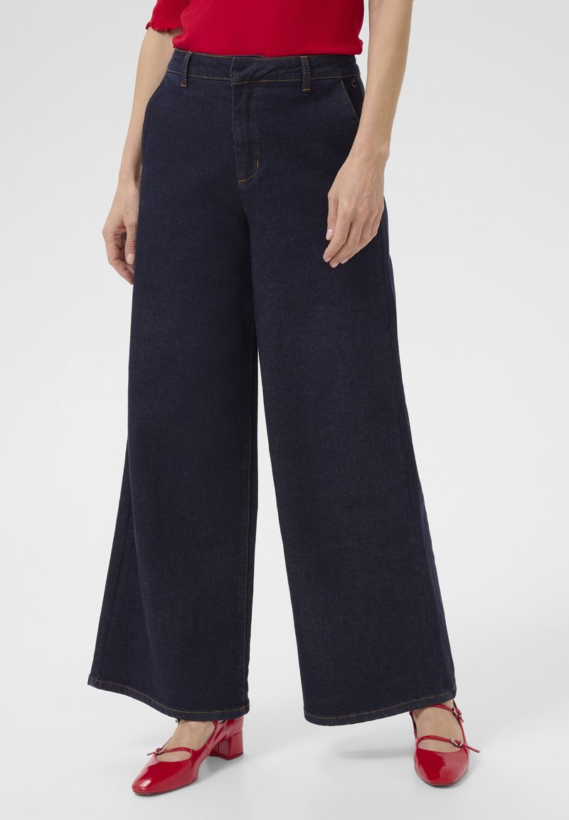 Löysät tummansiniset farkut korkealla vyötäröllä, joissa on näkyvät ompelusyksityiskohdat. Yhdistetty kiiltäviin punaisiin kenkiin.