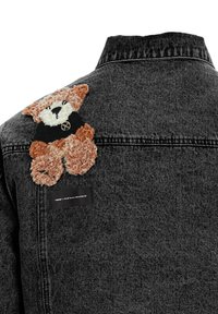 Chaqueta de mezclilla negra con un parche de oso texturizado y peludo en marrón y crema en la espalda, con una pequeña etiqueta negra cerca de la costura del hombro.