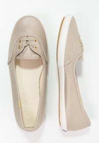 Chaussures slip-on en toile beige avec une surface texturée, des œillets dorés et une semelle blanche. Marquage sur la semelle intérieure ; design décontracté.