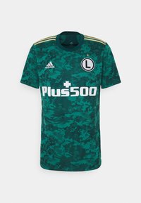 Zelený dres Adidas s kamuflážním vzorem, krátké rukávy, kulatý výstřih, bílá značka "Plus500" a klubový znak. Zlaté pruhy na ramenech.