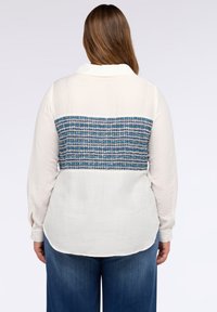 Camicia bianca a maniche lunghe con pannello posteriore in tessuto azzurro testurizzato, dotata di colletto e orlo curvo, abbinata a pantaloni in denim scuri.