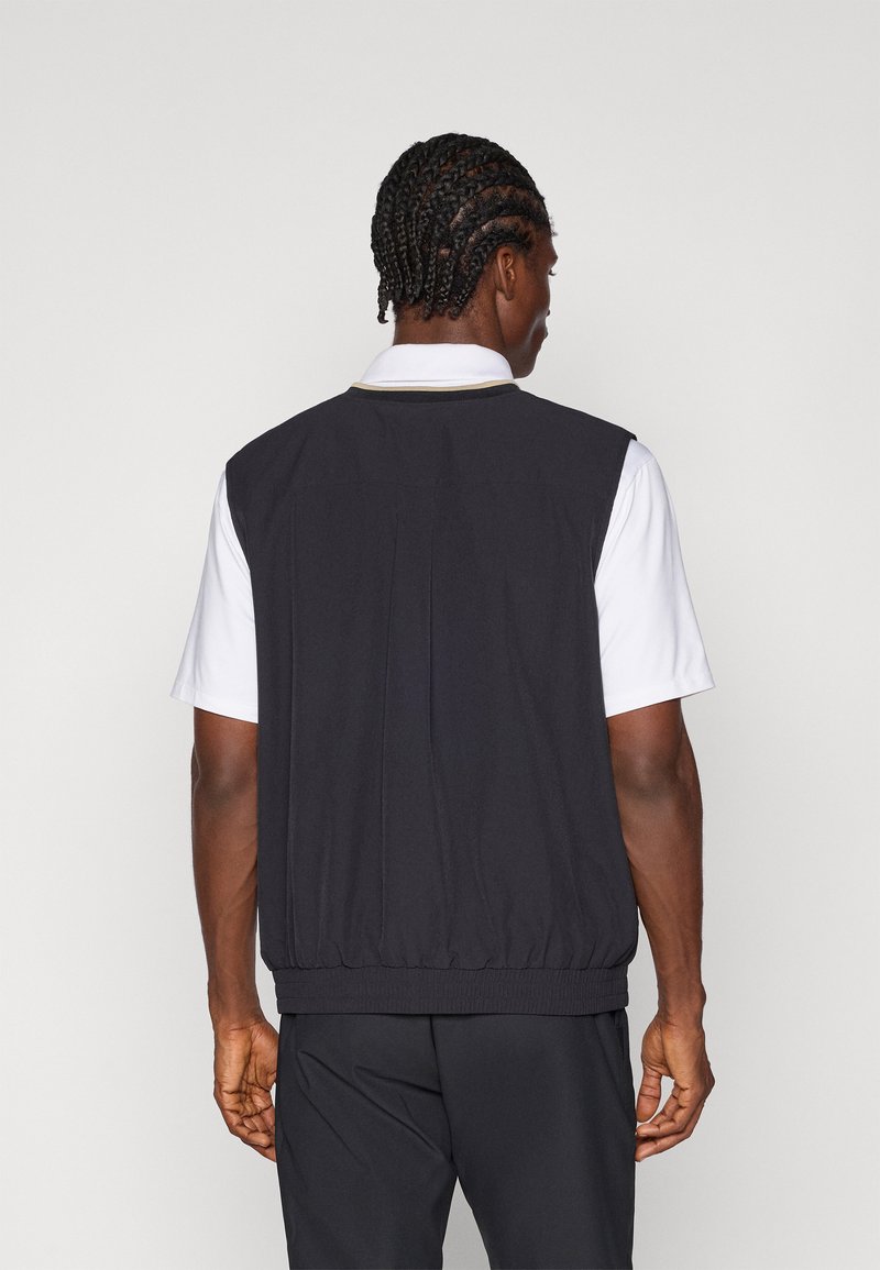 Veste sans manches noire avec ourlet élastique, présentant une texture lisse et un pli au dos. Portée sur une chemise blanche à col. Design simple et fonctionnel.