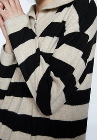 Felpa a righe beige e nere con colletto zip. Realizzata in tessuto morbido, presenta ampie righe orizzontali e polsini aderenti.