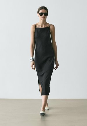 Donna che cammina con un vestito midi nero senza maniche con spacco laterale, ballerine bianche, bracciale a polsino argentato e occhiali da sole neri su sfondo neutro.