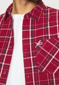 Rood geruite flanellen overhemd met witte en zwarte accenten, met een knoopsluiting en een borstzak met een logo aan de linkerkant. Gelaagd over een wit shirt.