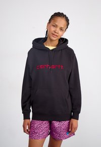 Black hoodie with red embroidered "carhartt." logo, featuring a kangaroo pocket and drawstring hood, paired with pink leopard print shorts.