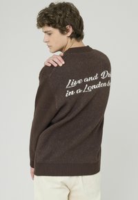 Pull en maille marron avec ourlet et poignets côtelés, comportant un texte blanc au dos qui dit "Vivre et Mourir à Londres" en écriture cursive.