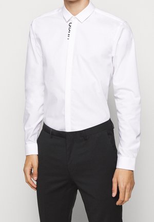 Mężczyzna ubrany w dopasowaną białą koszulę wizytową z długim rękawem, włożoną do czarnych eleganckich spodni, stojący z ramionami swobodnie opuszczonymi wzdłuż ciała.