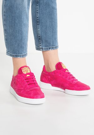 Fødder iført lyserøde Reebok-sneakers med hvide såler og gule logoer, parret med korte blå denimjeans på en hvid gulv.