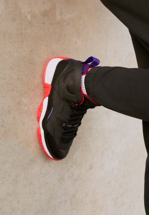 Schwarzer High-Top-Sneaker mit roter und weißer Sohle, getragen mit schwarzen Hosen auf Betonboden, seitlich und leicht von oben betrachtet.