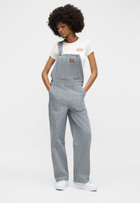 Νεαρή γυναίκα με σγουρά μαλλιά, που φοράει γυαλιά, λευκό μπλουζάκι, ριγέ ολόσωμη φόρμα και λευκά αθλητικά παπούτσια, στέκεται με τα χέρια στις τσέπες.