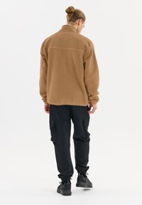 Veste en polaire beige avec un col haut, associée à un pantalon cargo noir. La veste présente une texture douce et une coupe décontractée. Des baskets sombres complètent l'ensemble.