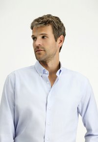 La camicia azzurra chiara ha un colletto e maniche lunghe. Tessuto liscio con una sottile consistenza e dettagli con bottoni standard.