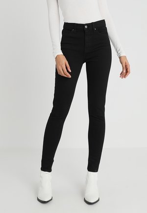 Femme portant un jean skinny noir taille haute, un haut blanc à manches longues et des bottines blanches sur un fond clair uni.