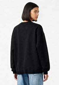 Vrouw met steil donker haar, gekleed in een losse zwarte trui en lichtblauwe jeans, licht zijwaarts gedraaid tegen een effen achtergrond.