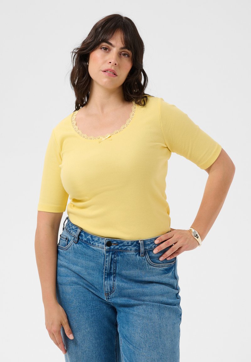 Femme aux cheveux foncés portant un haut jaune à manches courtes avec une bordure en dentelle et un jean bleu taille haute, debout, une main sur la hanche.