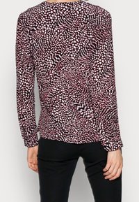 Vue arrière d'une personne portant un blouse à manches longues avec un motif abstrait noir, blanc et rose et un pantalon noir, se tenant contre un fond uni.