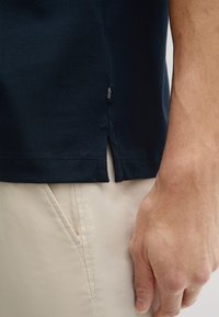 Camiseta azul marino de algodón, con una abertura lateral y una pequeña etiqueta de la marca en el dobladillo. Mano en primer plano, combinada con pantalones beige claros.