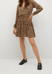 Robe en imprimé léopard couleur beige avec des manches longues et une taille smockée. Associée à des mocassins noirs avec des accents dorés. Texture de tissu douce.