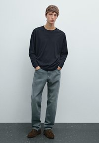 Maglietta nera a maniche lunghe, jeans grigi chiari, scarpe marroni. Vestibilità casual, tessuto morbido e design semplice. Pose in piedi su uno sfondo semplice.