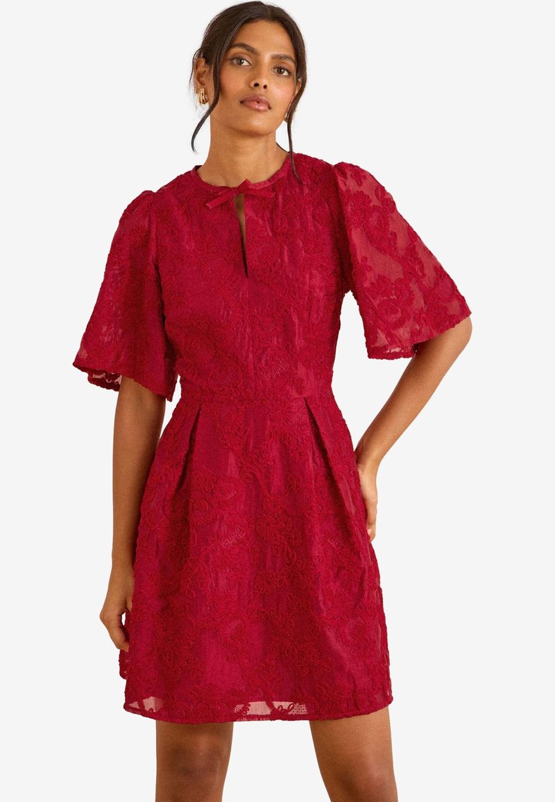 Mujer con vestido corto de encaje rojo con mangas amplias y pequeño escote en forma de ‘ojo de cerradura’ atado con un lazo, posando con una mano en la cadera.