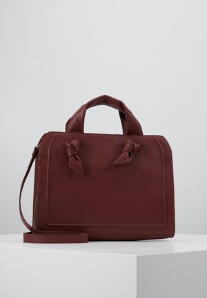 Sac à main en cuir rouge foncé avec deux poignées nouées et une bandoulière amovible sur une surface blanche contre un fond gris.