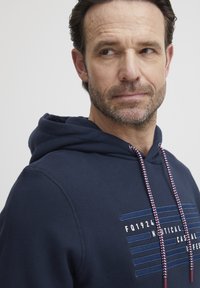 Sudadera azul marino hecha de tela suave. Cuenta con una capucha, cordones con un patrón y texto bordado con rayas horizontales en la parte frontal.