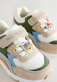 Zapatillas infantiles con gamuza beige, malla blanca, detalles en verde y azul, correas de velcro con la etiqueta "Paw Patrol" y placas de personajes en los cordones blancos.