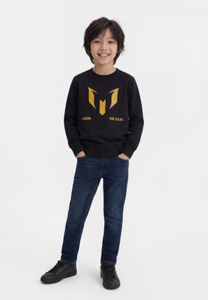 Uśmiechnięty chłopiec o czarnych włosach stoi z rękami w kieszeniach, ubrany w czarną bluzę z kapturem z złotym logo i napisem, niebieskie dżinsy oraz czarne trampki.