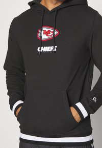 Hoodie noir avec un logo brodé des Kansas City Chiefs en rouge et blanc, une poche frontale et des rayures blanches contrastantes aux poignets et à l'ourlet.