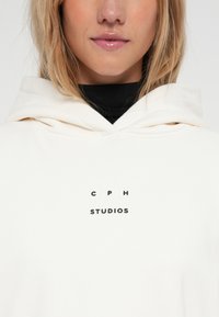 Biela mikina s kapucňou a predným vreckom, na ktorej je čierny potlačený logo so slovami "C P H STUDIOS." Materiál vyzerá byť mäkký, s voľným strihom.