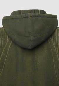 Giacca verde oliva con cappuccio, con cuciture e dettagli delle cuciture gialli visibili su uno sfondo chiaro e neutro.