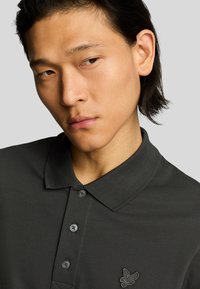 Polo di colore grigio scuro con colletto, tre bottoni e tessuto testurizzato. Logo ricamato ben visibile sul petto.