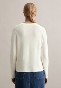 Cremfarbener Strickpullover mit langen Ärmeln, rundem Ausschnitt, gerippter Textur und geradem Saum. Kombiniert mit einem dunkelblauen Denimrock mit Taschen.