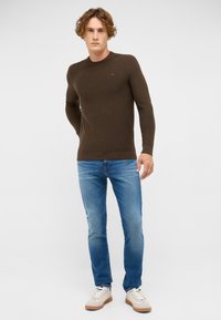 Brązowy sweter prążkowany z długimi rękawami, połączony z jasnoniebieskimi dżinsami i kremowymi sneakersami. Sweter ma subtelne logo na piersi.