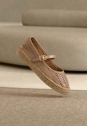 Pantof bej perforat, cu talpă plată, vârf rotunjit, curea reglabilă și talpă din iută împletită, suspendat deasupra unei suprafețe texturate.
