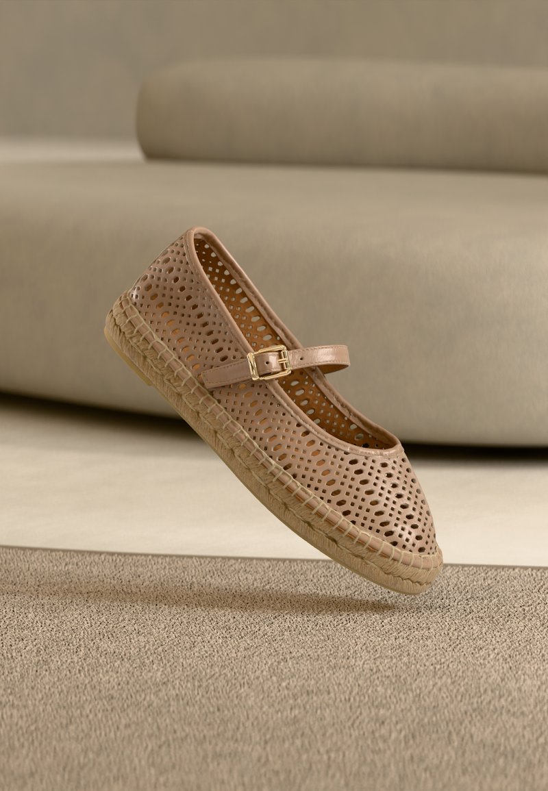 Chaussure plate beige perforée avec un bout arrondi, une bride ajustable et une semelle en jute tissée, suspendue au-dessus d'une surface texturée.