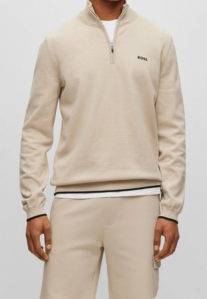 Beige pullover sweater with a quarter-zip collar, ribbed cuffs, and hem. Features a small black logo on the chest and is made from soft fabric.