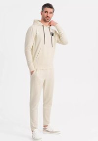 Cremefarbener Hoodie und passende Jogginghose aus weichem Stoff, mit einer Vordertasche, schwarzen Zugbändern und einem Logo auf der Brust.