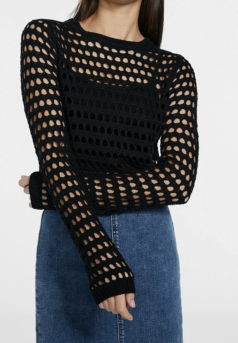 Femme portant un pull en crochet noir à manches longues avec de grands trous par-dessus un top noir et une jupe en jean bleue.