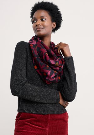 Kvinde med kort krøllet hår iført sort prikket cardigan, farverig mønstret rød og lilla tørklæde og røde cordbukser, kigger sidelæns.