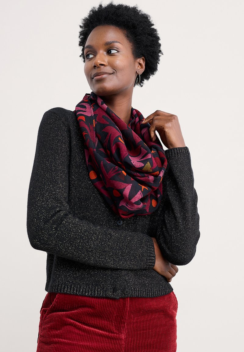 Vrouw met kort krullend haar draagt een zwart gespikkeld vest, een kleurrijke gestreepte rode en paarse sjaal, en rode ribfluweel broek, terwijl ze opzij kijkt.