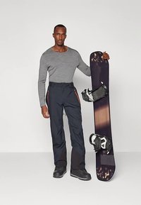 Άνδρας φορώντας γκρι μπλουζάκι με μακριά μανίκια και μαύρο παντελόνι χιονιού κρατάει κατακόρυφα μαύρη σανίδα snowboard με δεσίματα μπροστά σε άσπρο φόντο.