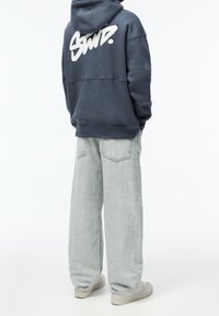 Námořnická mikina s kapucí a bílým logem na zádech, zkombinovaná se světlými šedými širokými jeansy a světlými teniskami, z pohledu zezadu.