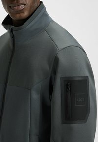 Grå zip-up jacka med hög krage, strukturerad tyg och en bröstficka med dragkedja och en svart logotypapplikation.