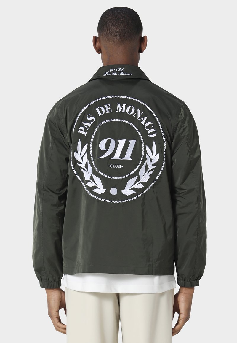 Mężczyzna w ciemnozielonej kurtce z białym napisem "PAS DE MONACO 911 CLUB" i wzorem laurowego wieńca na plecach, stojący na białym tle.