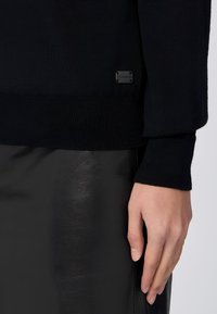 Černý pletený svetr s žebrovanými manžetami, na boku s malou černou etiketou. V kombinaci s lesklými černými koženými kalhotami.