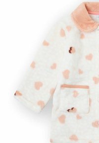 Chaqueta borrosa color crema con patrones de corazones rosas, un cuello suave y un bolsillo delantero con borde melocotón. Textura acogedora y corte holgado.