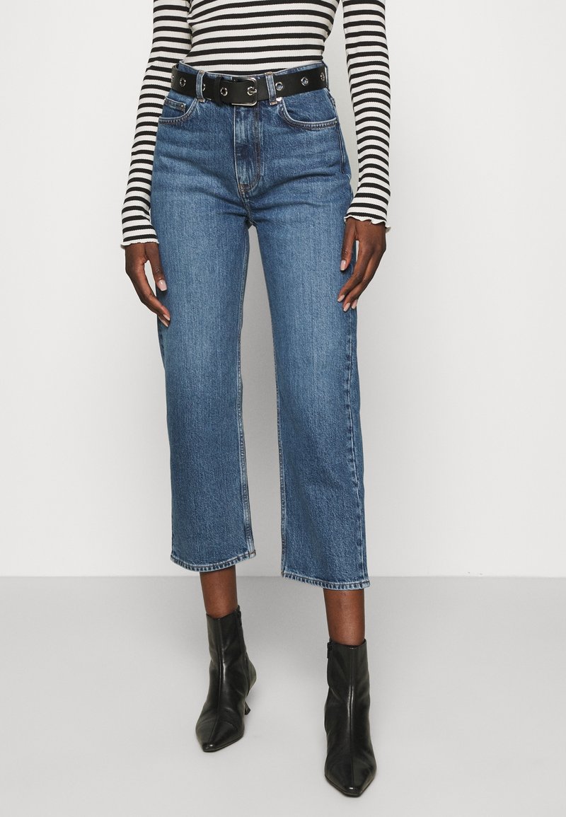 Höga blå denimcroppade jeans med rak passform, slitna fållar och svart bälte med metallbeslag. Randig långärmad topp.