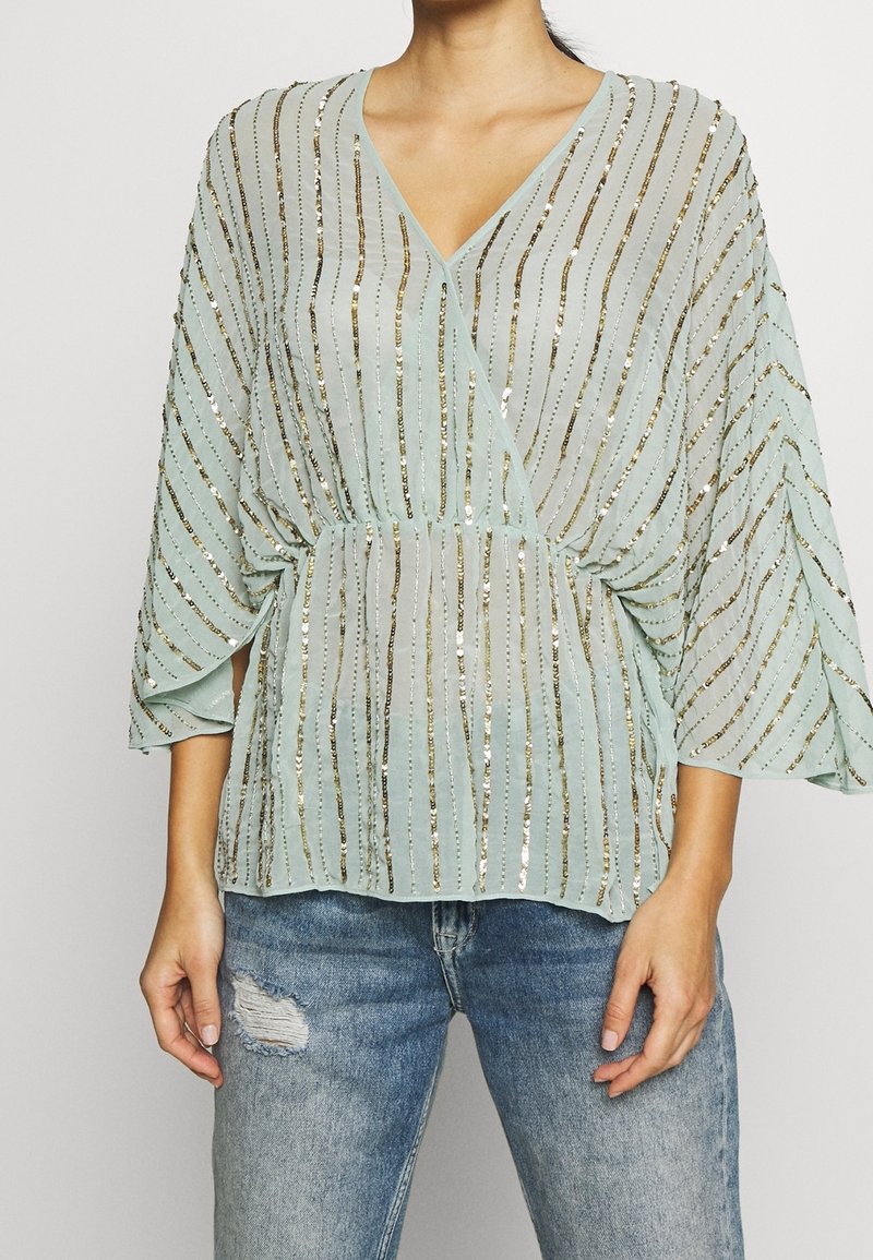 Blouse bleu clair à col en V avec des rayures verticales en sequins dorés et des manches larges, associée à un jean bleu clair déchiré.